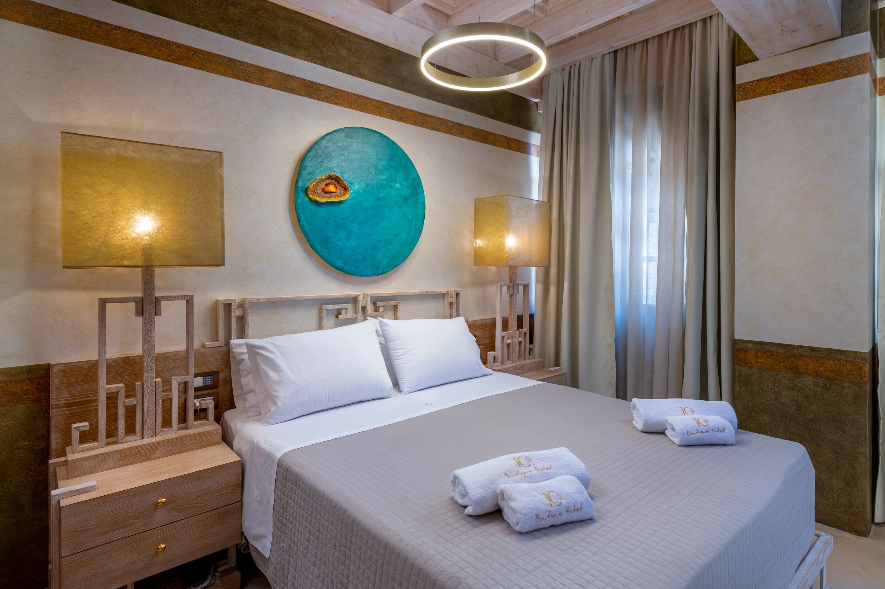 A hotel room with a large bed, two bedside tables, a modern wall art piece, and a large window with curtains.
