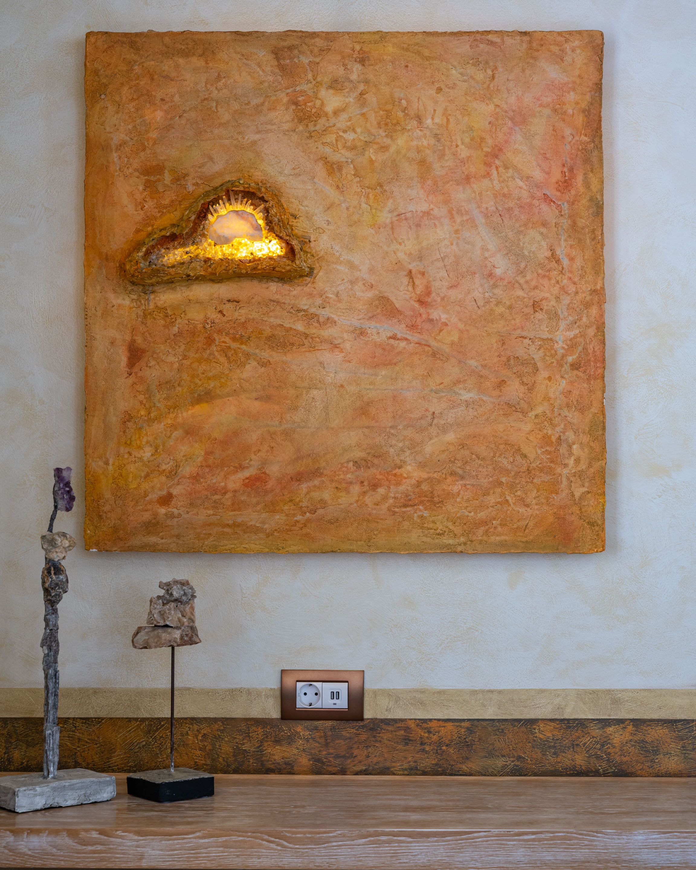 A large abstract painting with a warm color palette on a textured wall. Below the painting, there is a power outlet and a small sculpture on a wooden base.