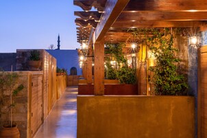 Área externa coberta com vigas de madeira, plantas em vasos e luminárias fixadas na parede.