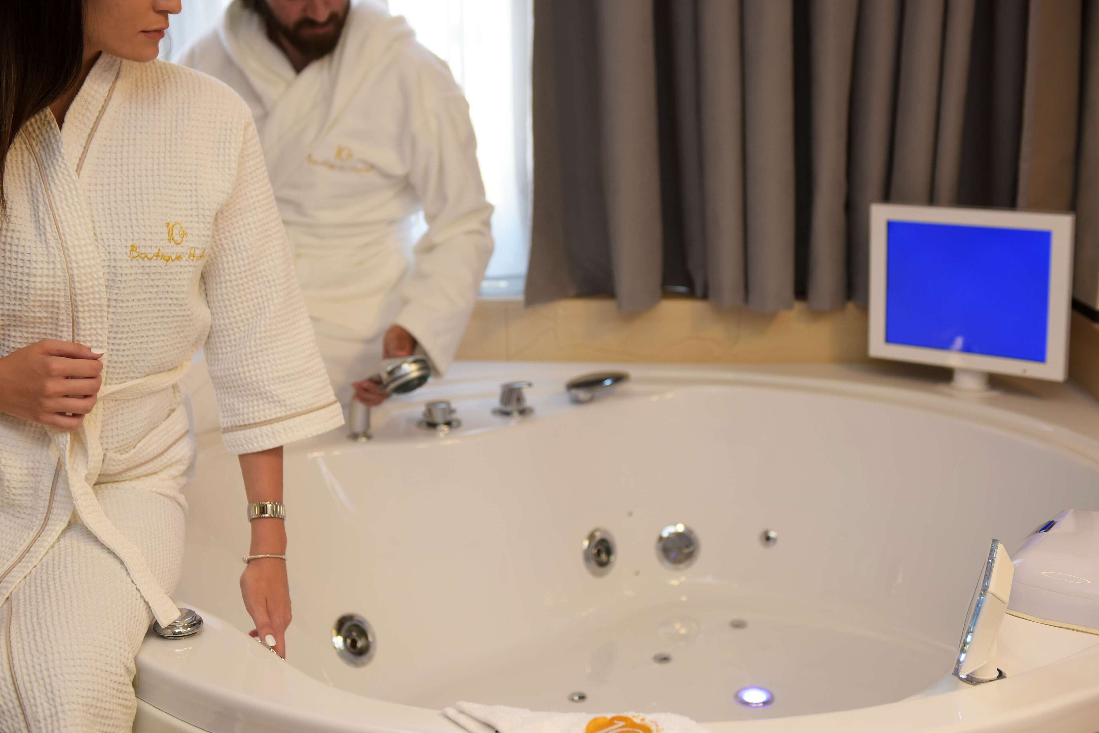A large bathtub with multiple faucets and a handheld showerhead, a person in a robe, and a television screen in the background.