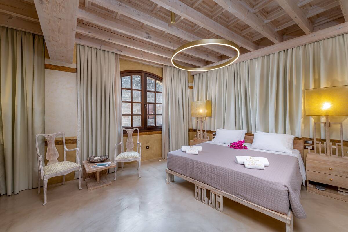 Une chambre avec un plafond en bois, une grande fenêtre, un lit avec des oreillers et une couverture, deux chaises, une table de chevet et une lampe.