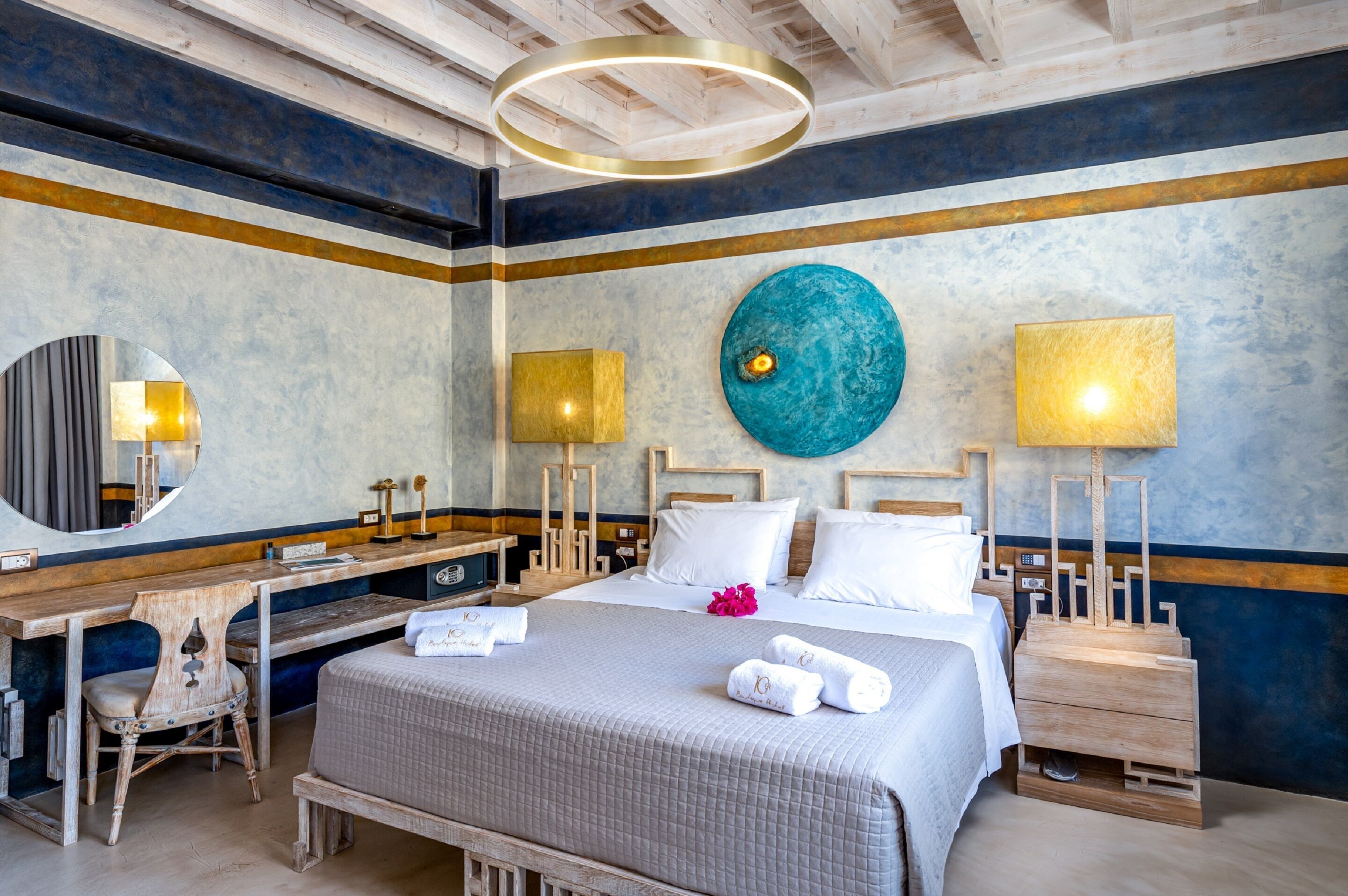A hotel room with a large bed, bedside tables, a desk, a chair, a mirror, and a circular wall decoration.