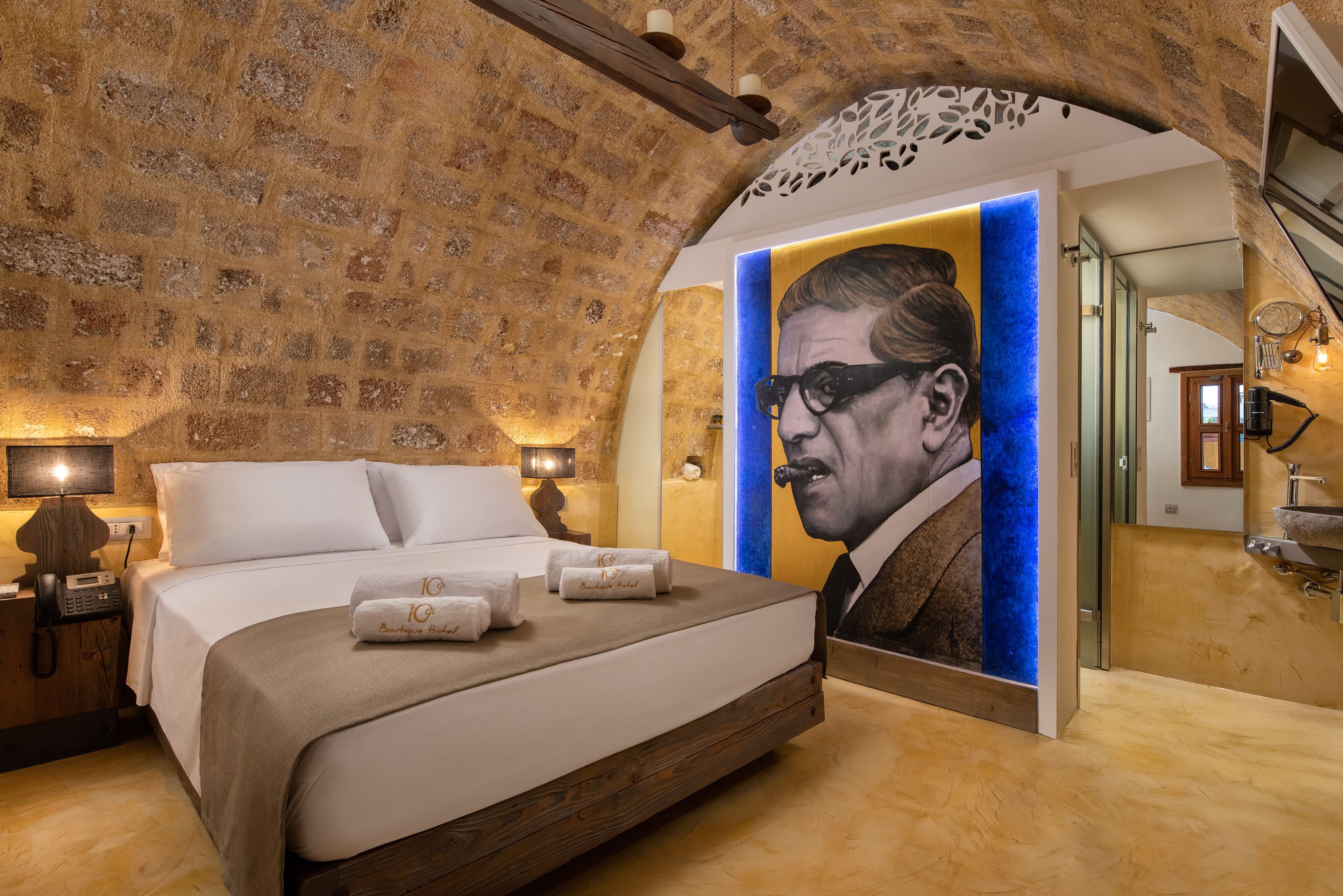 Una habitación de hotel con una cama grande, cabecera de madera, un mural de un retrato y una pared de piedra.