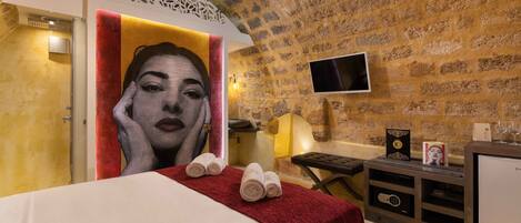 A hotel room with a stone wall, a bed with a red blanket, a TV, and a massage table.