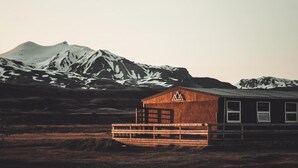 Une cabane en bois surmontée d’un toit en métal, entourée d’une terrasse en bois, le tout se dressant sur un fond de montagnes enneigées.