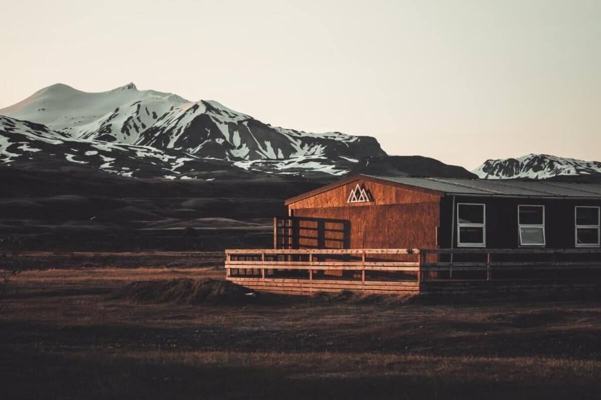 A wooden cabin with a metal roof, surrounded by a wooden deck, set against a backdrop of snow-capped mountains.