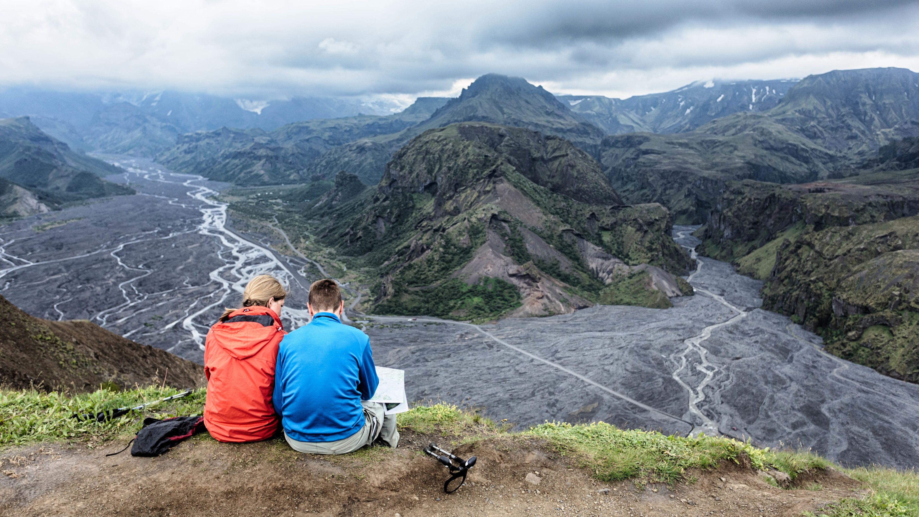 Two people sitting on a hillside overlooking a valley with a river and mountains.