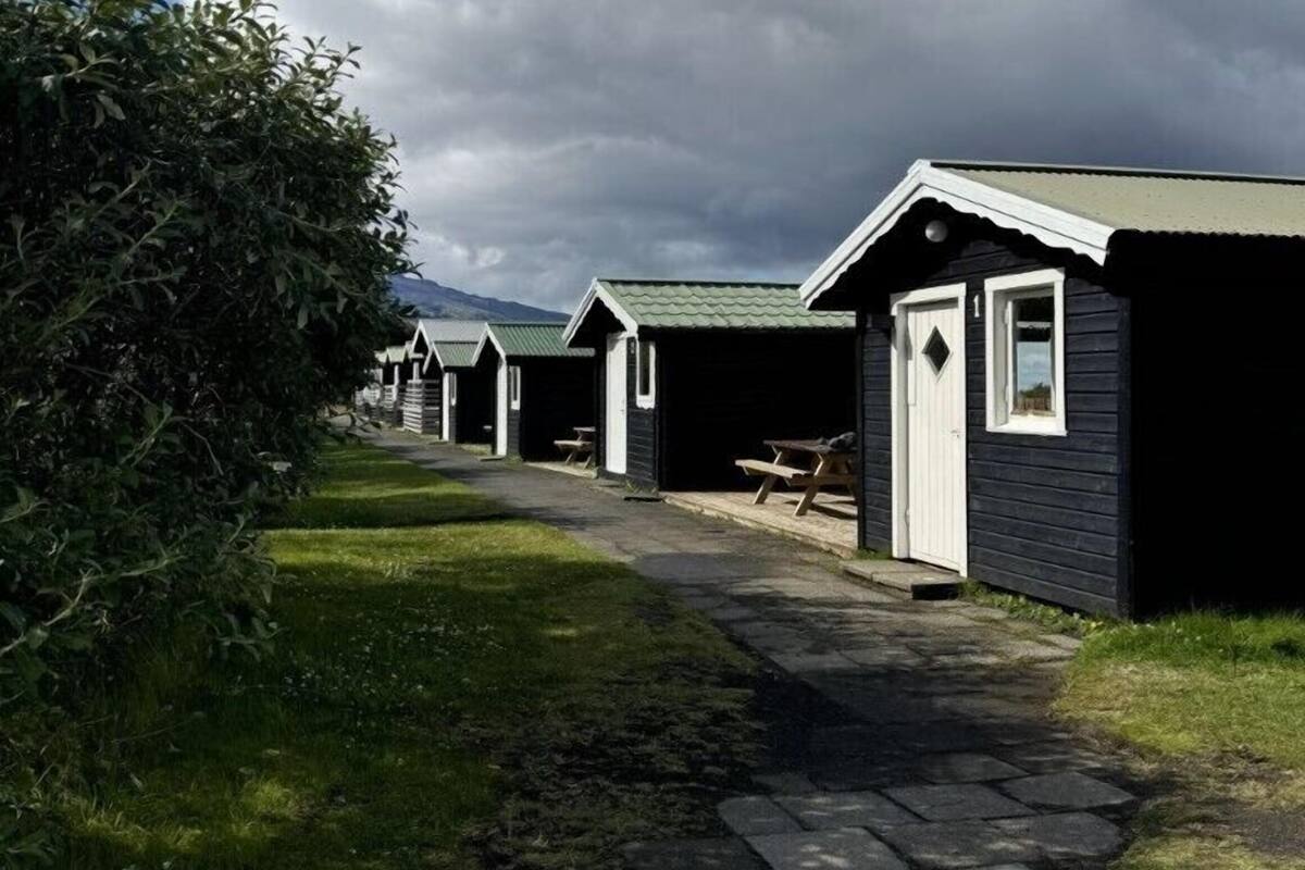 A row of small, dark-colored cabins with a stone pathway leading to them, surrounded by greenery and under a cloudy sky.