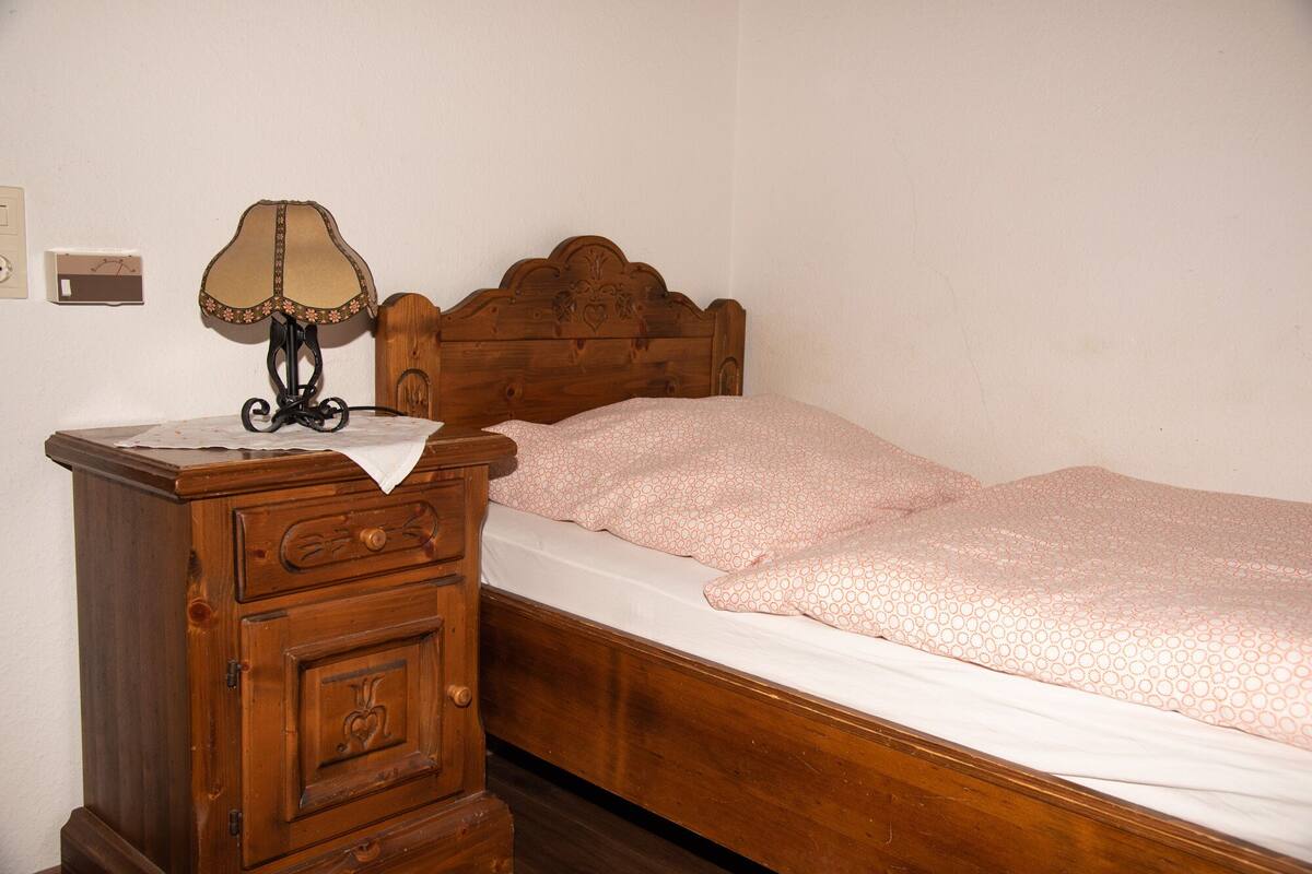 A wooden bed with a headboard, a wooden nightstand with a lamp, and a wall-mounted light switch.