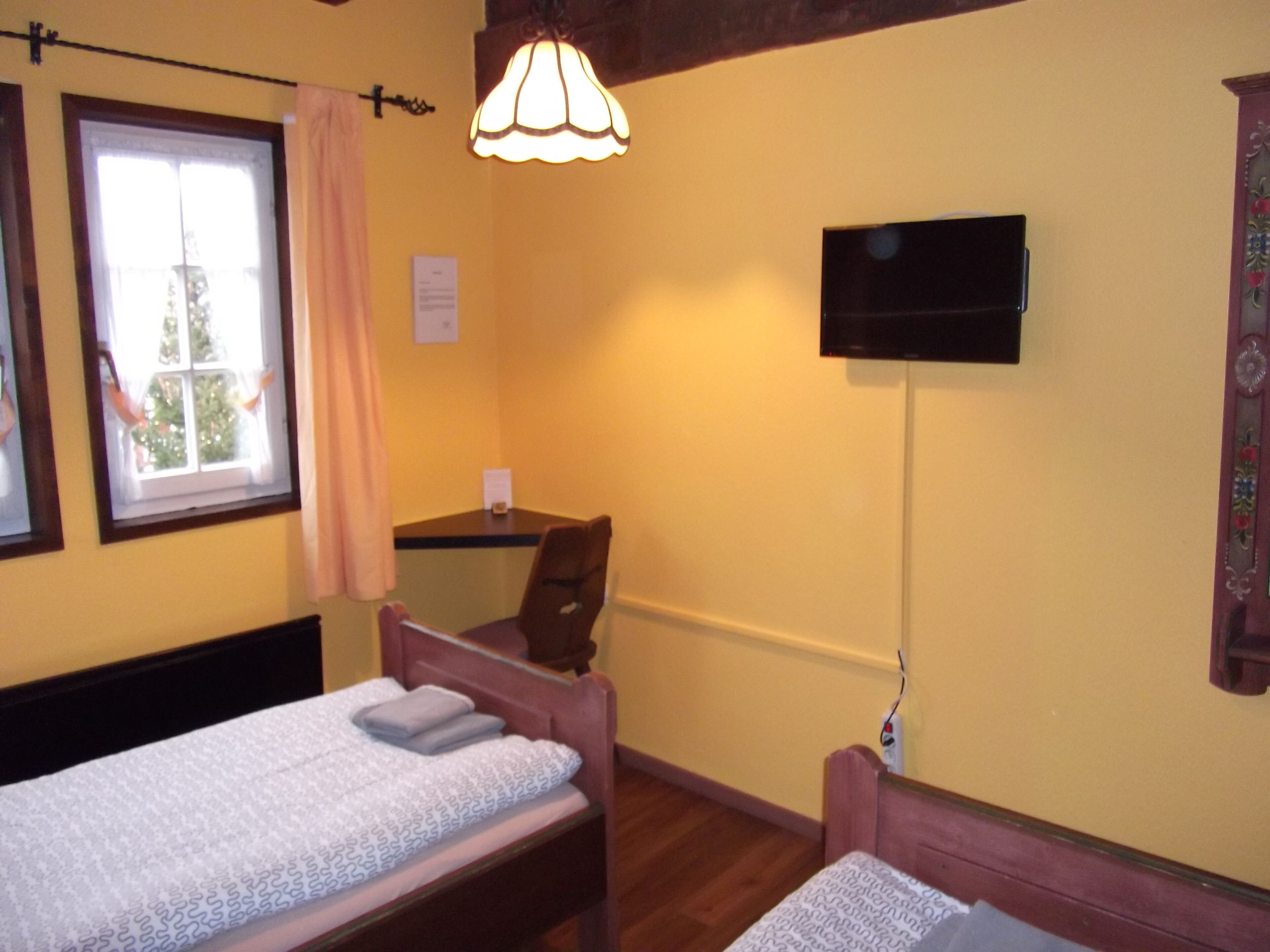 A small, cozy room with two single beds, a desk, a chair, a television, and a window with a curtain.
