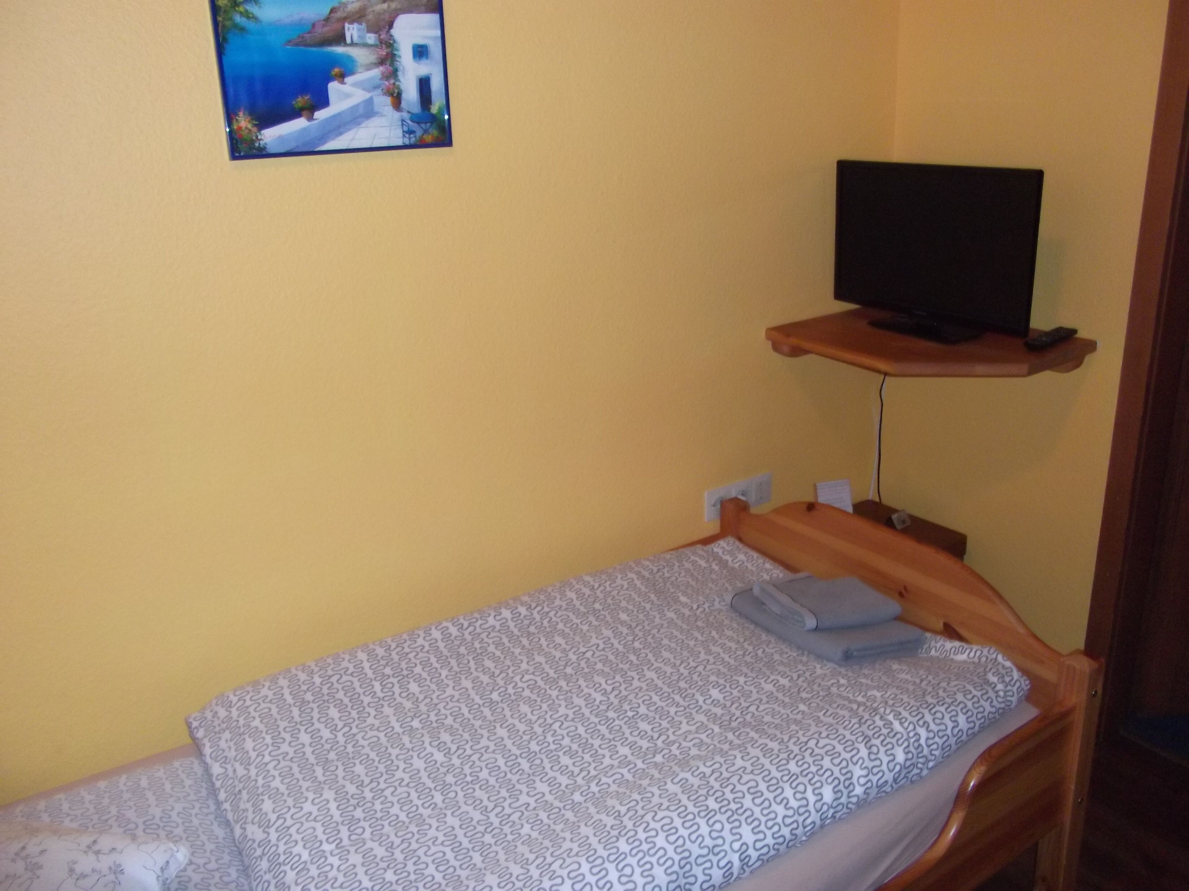 Ein kleines Zimmer mit einem Einzelbett, einem Fernseher auf einer Holzablage und einem Gemälde, das eine Küstenlandschaft zeigt.