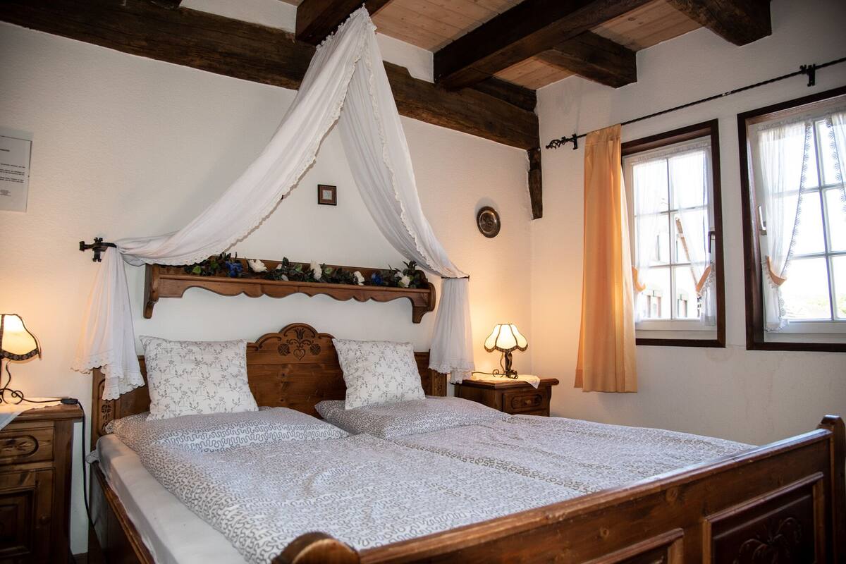 A bedroom with a wooden bed, a canopy, two bedside tables with lamps, and a window with curtains.