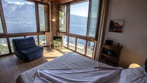 A bedroom with a large bed, a chair, and a view of a lake and mountains through large windows. - Hotel e Appartamenti al Prà de la Fam (Tignale)