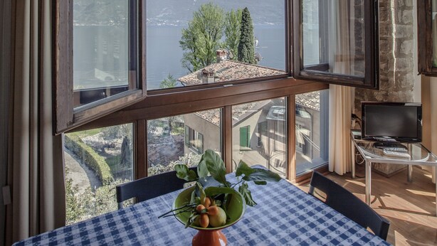 A room with a large window offering a view of mountains and a lake. There is a table with a checkered tablecloth and a potted plant. - Hotel e Appartamenti al Prà de la Fam (Tignale)