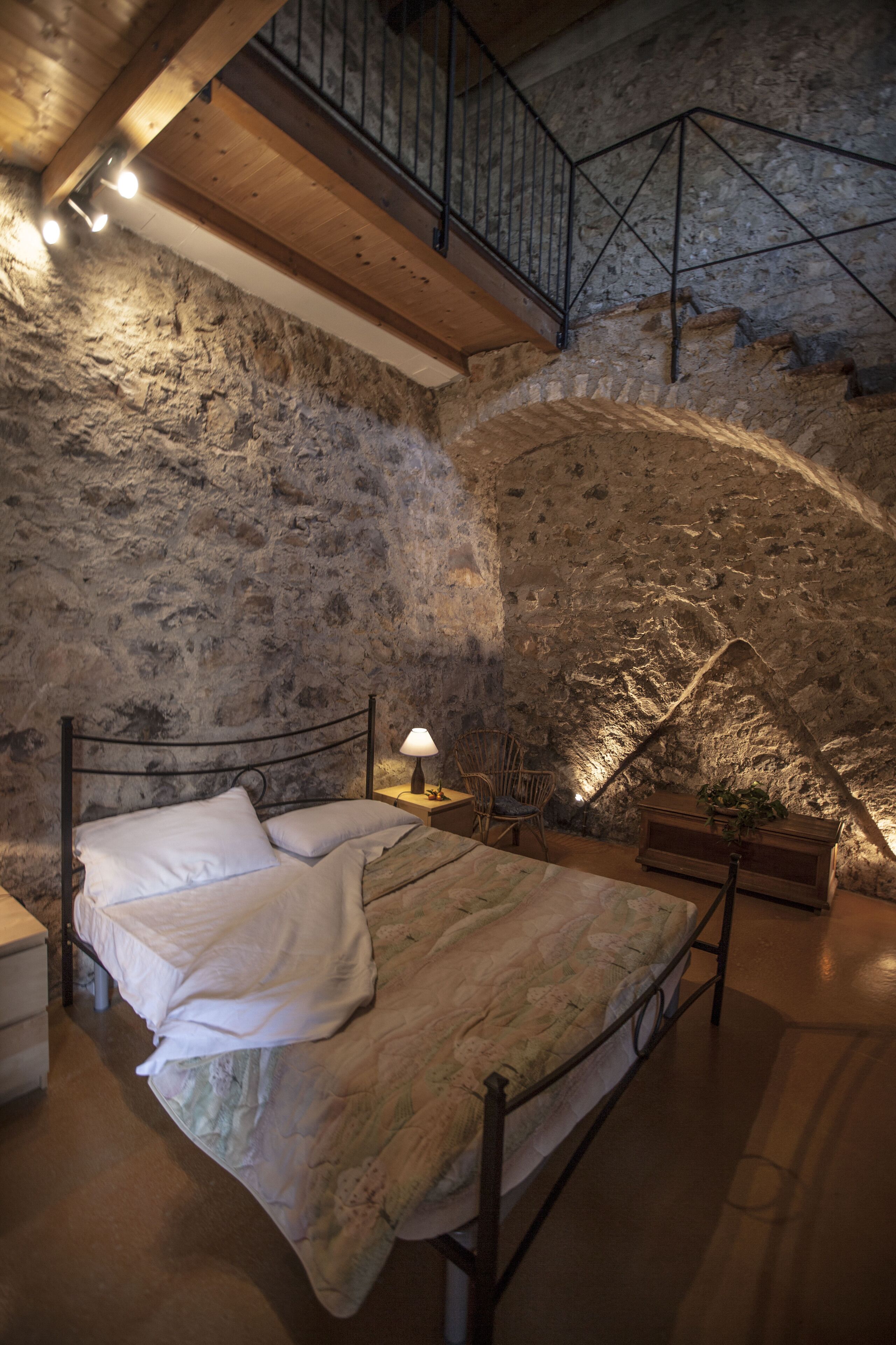 A bedroom with a stone wall, a wooden bed, a nightstand, a chair, and a staircase.