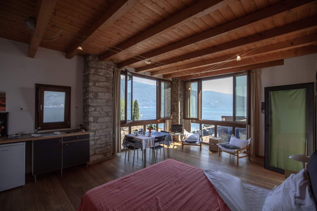 Un dormitorio con una cama grande, vigas de techo de madera, pared de piedra, un escritorio con ordenador y vistas a montañas y al agua.
