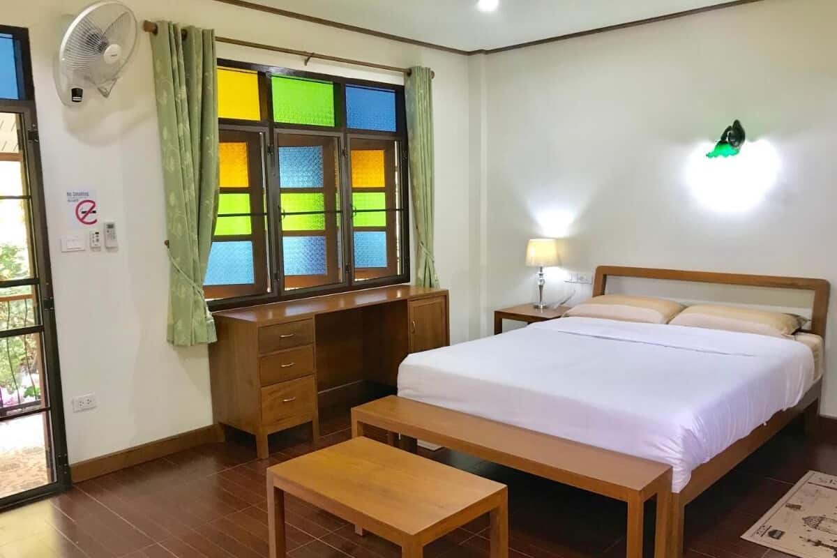 A bedroom with a bed, bedside table, wooden desk, and a window with colorful panes.