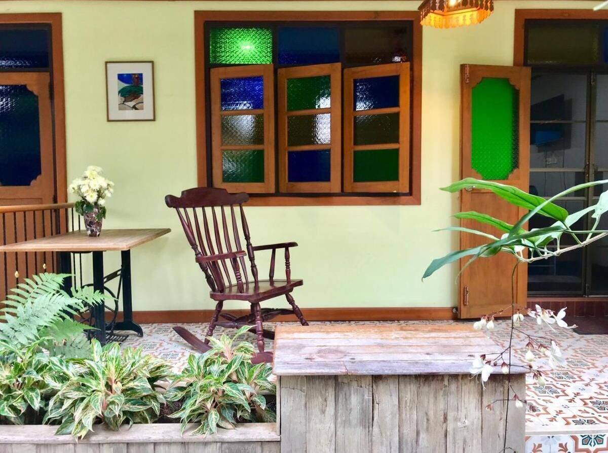A wooden chair near a window with stained glass panes, a table with a vase of flowers, and various potted plants.