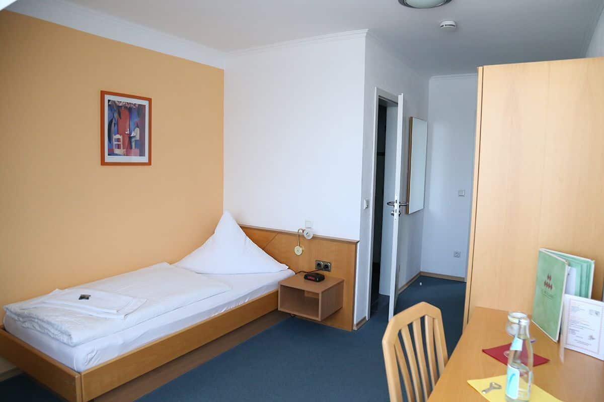 Ein Einzelbett mit Holz Kopfteil, ein Nachttisch mit Lampe, ein Stuhl, ein Tisch mit Flasche und Papier, sowie ein gerahmtes Bild an der Wand.