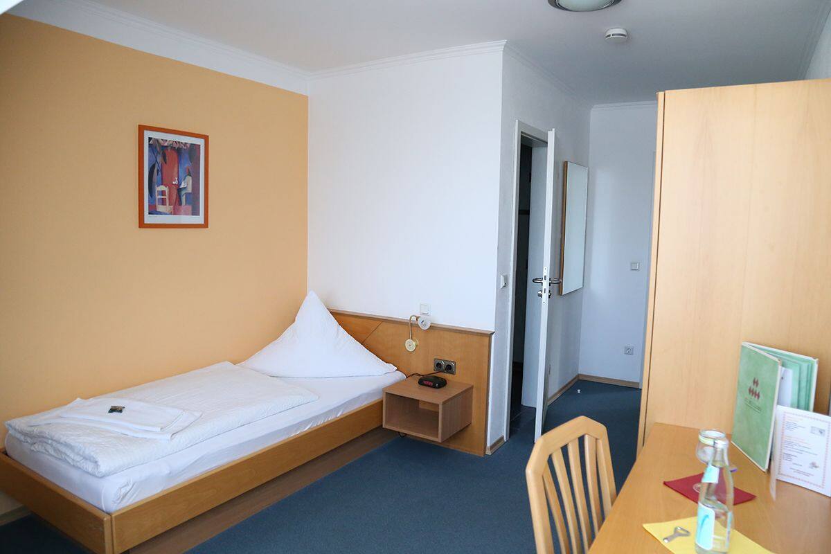 Ein Einzelbett mit Holz-Kopfteil, ein Nachttisch mit Lampe, eine kleine Ablage und ein gerahmtes Bild an der Wand.