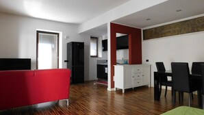 A modern living room with a red sofa, a dining area with black chairs, a white cabinet, and a kitchen in the background. - Garden Apartment (Verona)