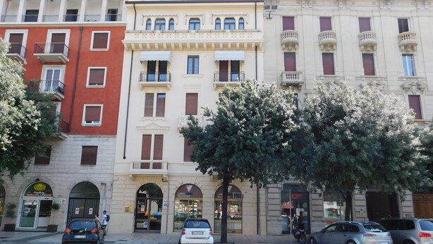 A row of multi-story buildings with various storefronts, including a pharmacy and a bank. - Garden Apartment (Verona)