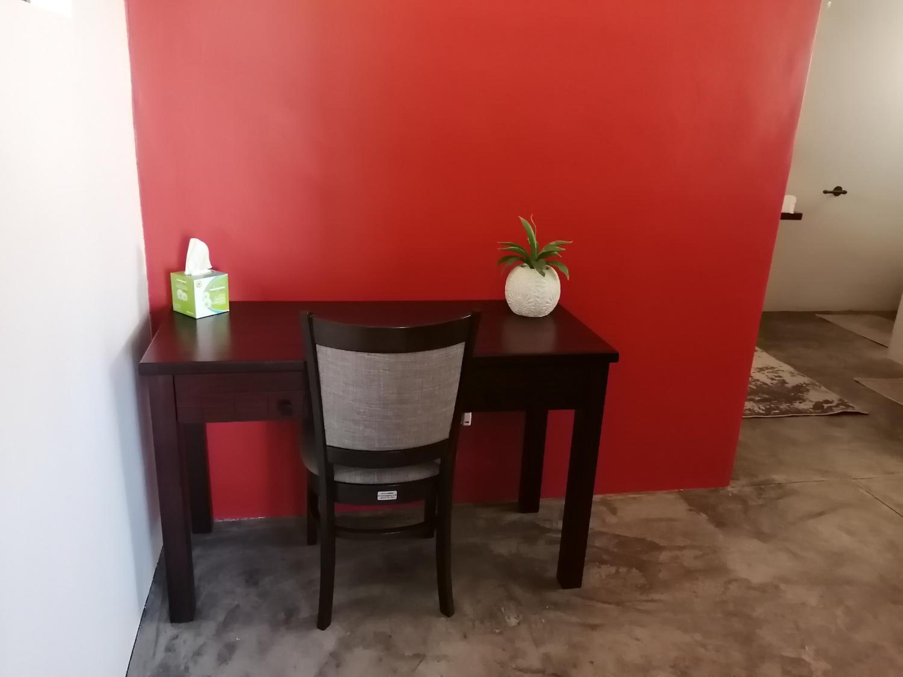 A small, dark wooden desk with a chair, a tissue box, and a potted plant against a red wall.