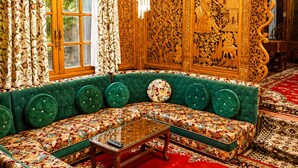 A room with a green upholstered sofa, a glass coffee table, ornate wooden carvings, and a chandelier. - De-Laila Houseboats (Srinagar)