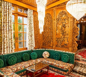 A room with a green upholstered sofa, a glass coffee table, ornate wooden carvings, and a chandelier. - De-Laila Houseboats (Srinagar)