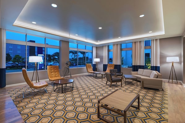 A spacious hotel lobby with a geometric patterned carpet, leather armchairs, a sofa, and a dining table. Large windows offer views of the outside.