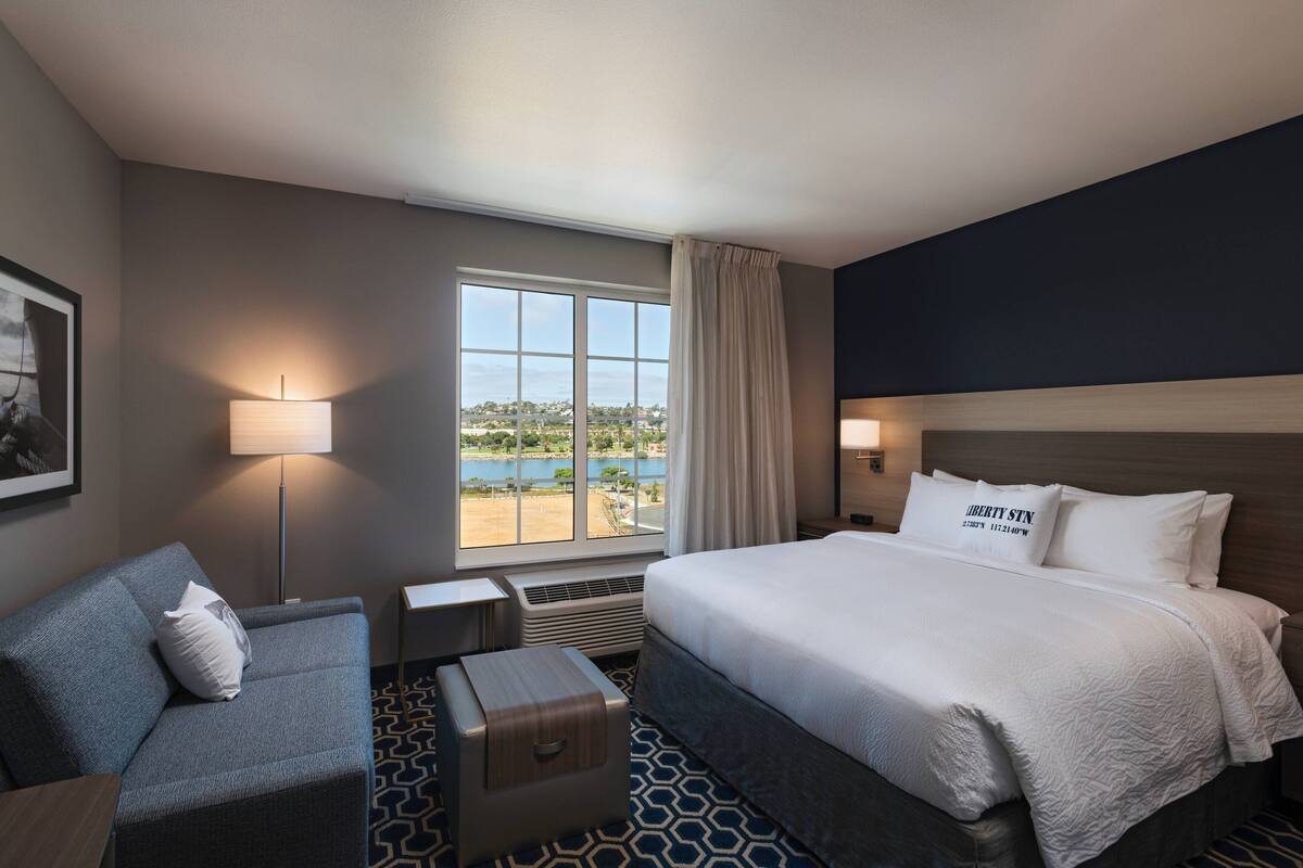 A hotel room with a bed, a sofa, a lamp, a window with a view of a beach, and a small table.