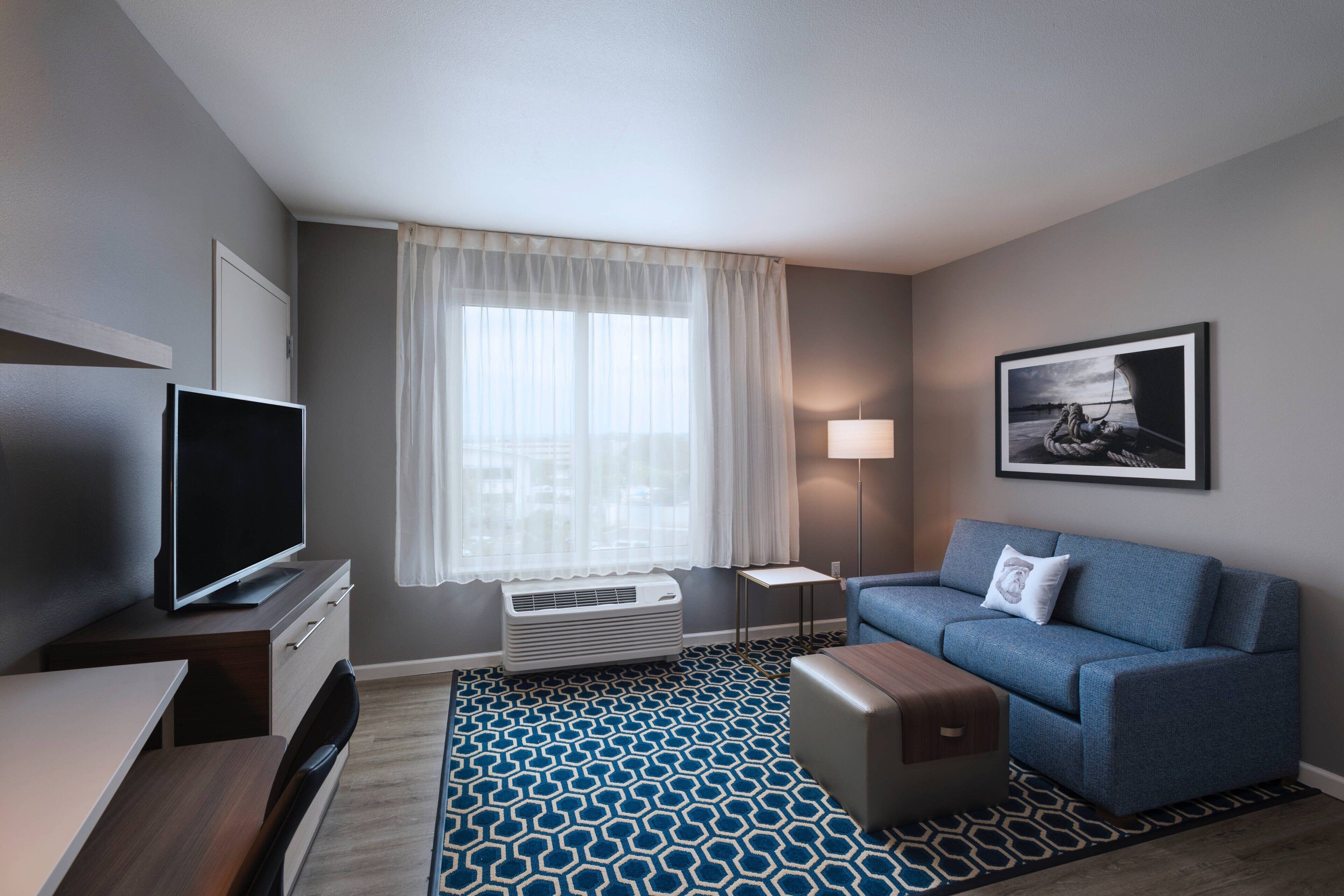 Ein modernes Hotelzimmer mit Flachbildfernseher, einem blauen Sofa, einem Holztisch, einem gemusterten Teppich, einem großen Fenster mit transparenten Vorhängen, einer Stehlampe und einem gerahmten Bild an der Wand.