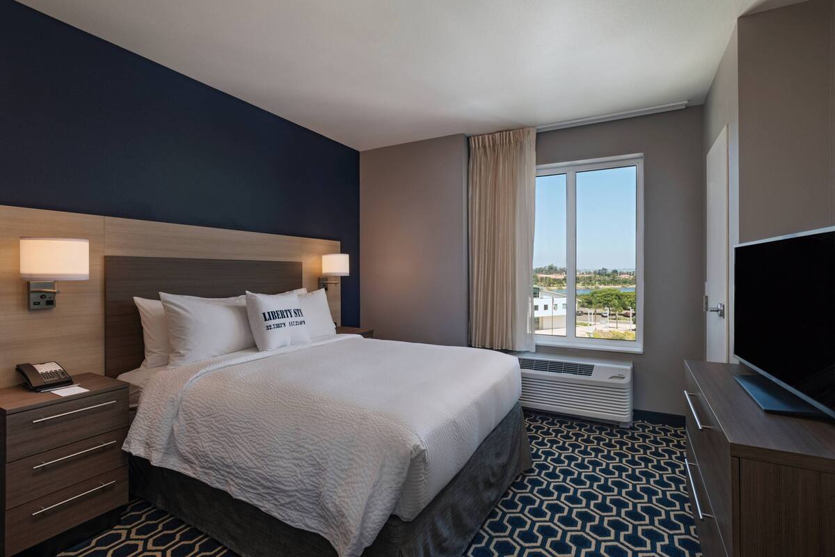 A hotel room with a bed, bedside lamps, a TV, a window with curtains, and a view of the outside.