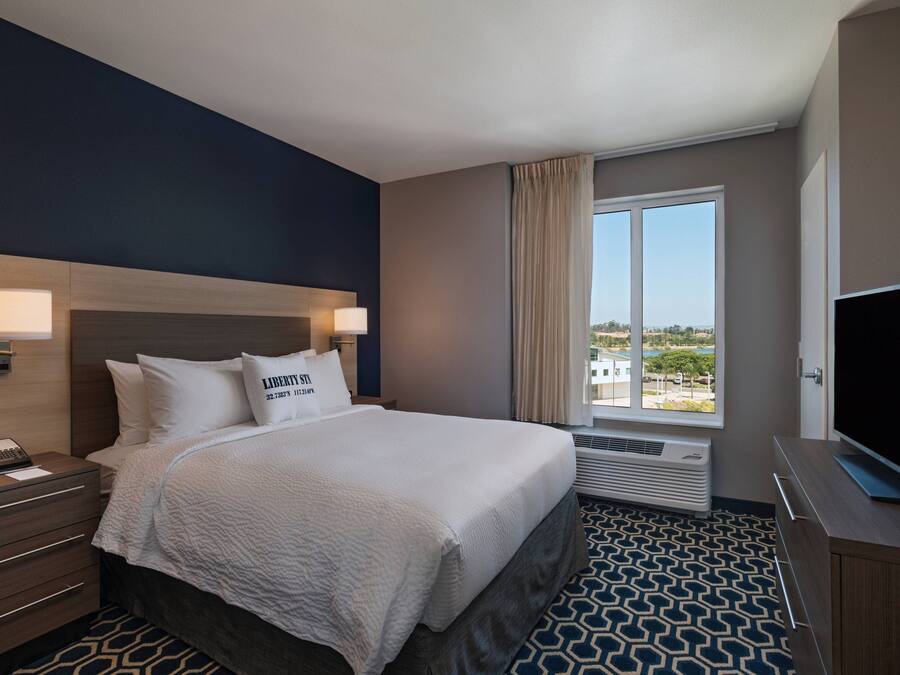 A hotel room with a bed, bedside lamps, a TV, a window with curtains, and a view of the outside.