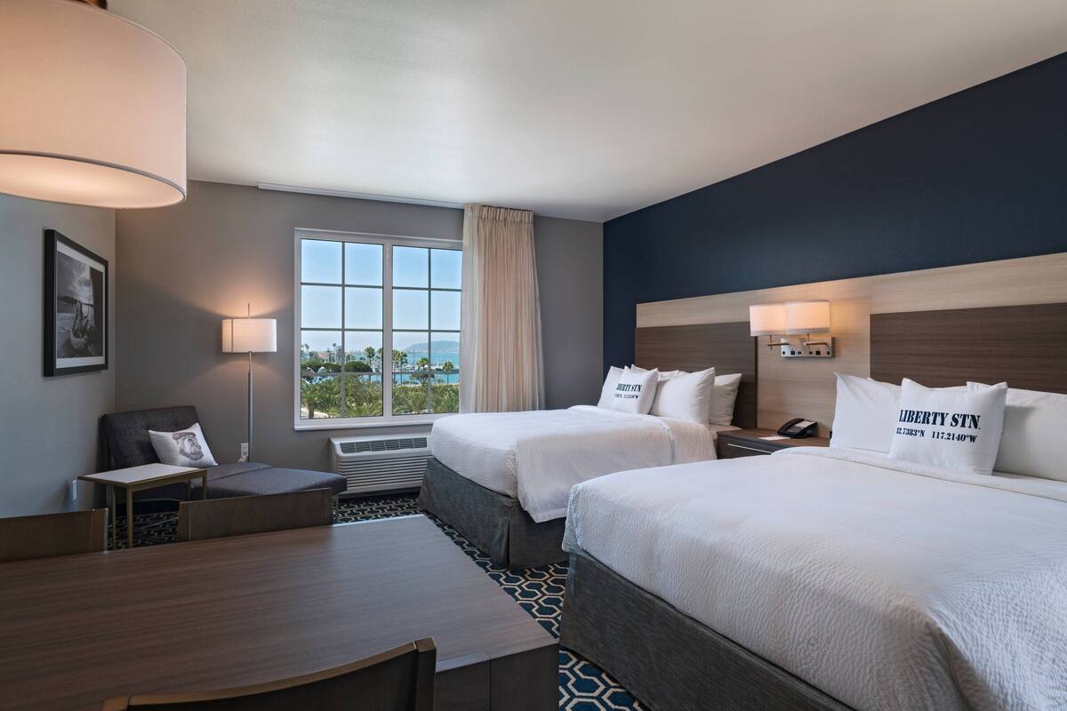 A hotel room with two beds, a desk, a chair, a window with a view, and a lamp.