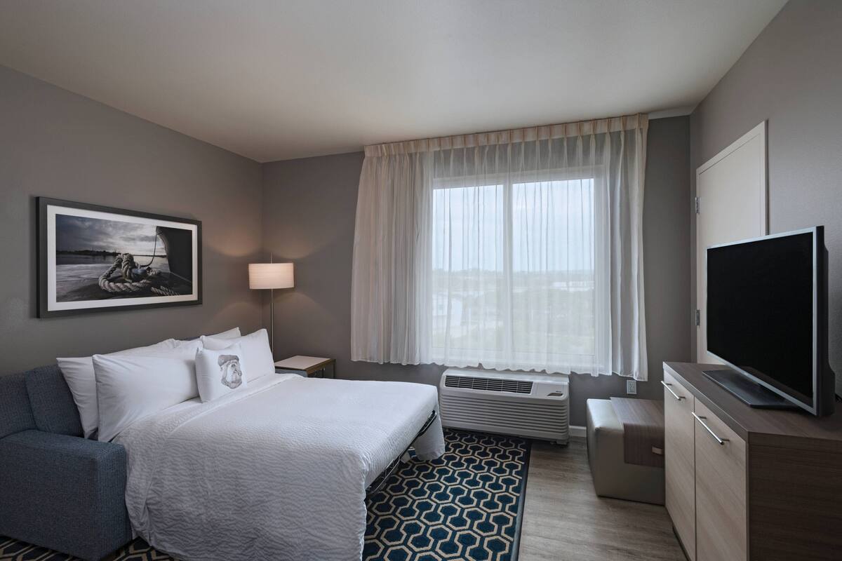 A hotel room with a bed, a TV, a desk, a lamp, and a window with curtains.