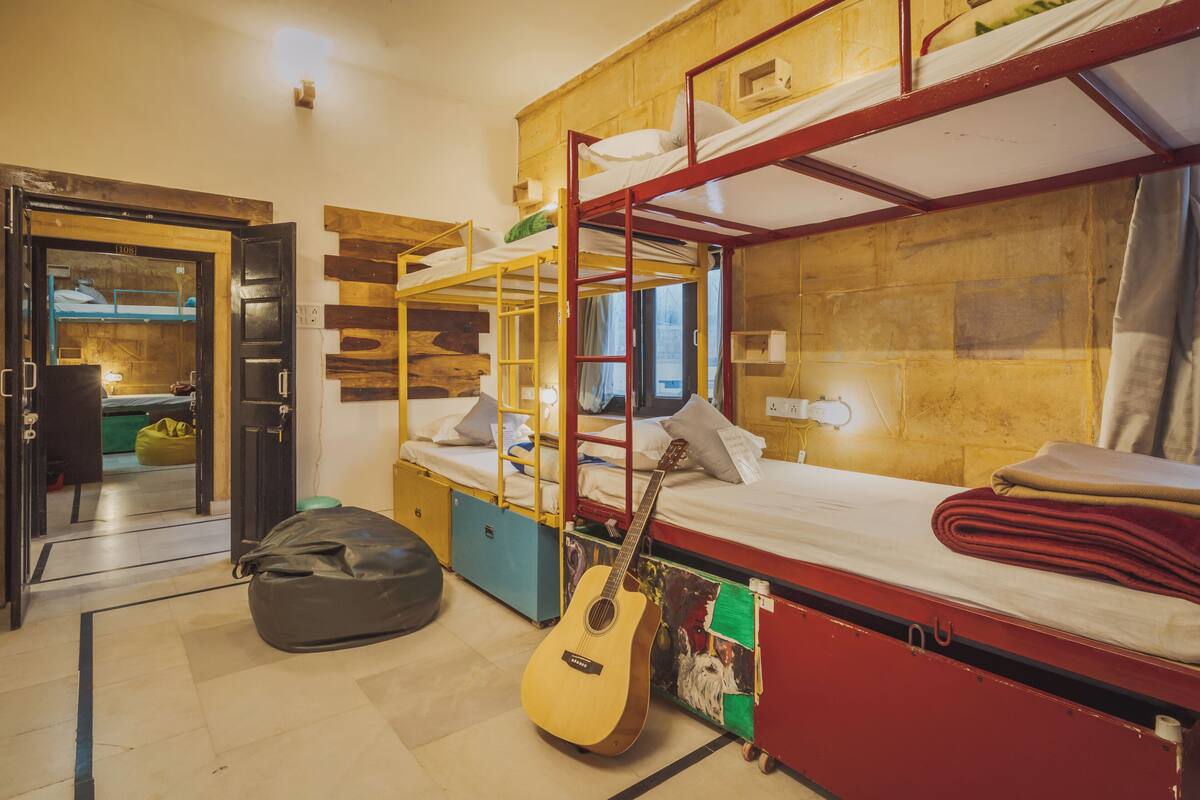 Quarto em dormitório com beliches, um violão, um puff e uma pequena cozinha.