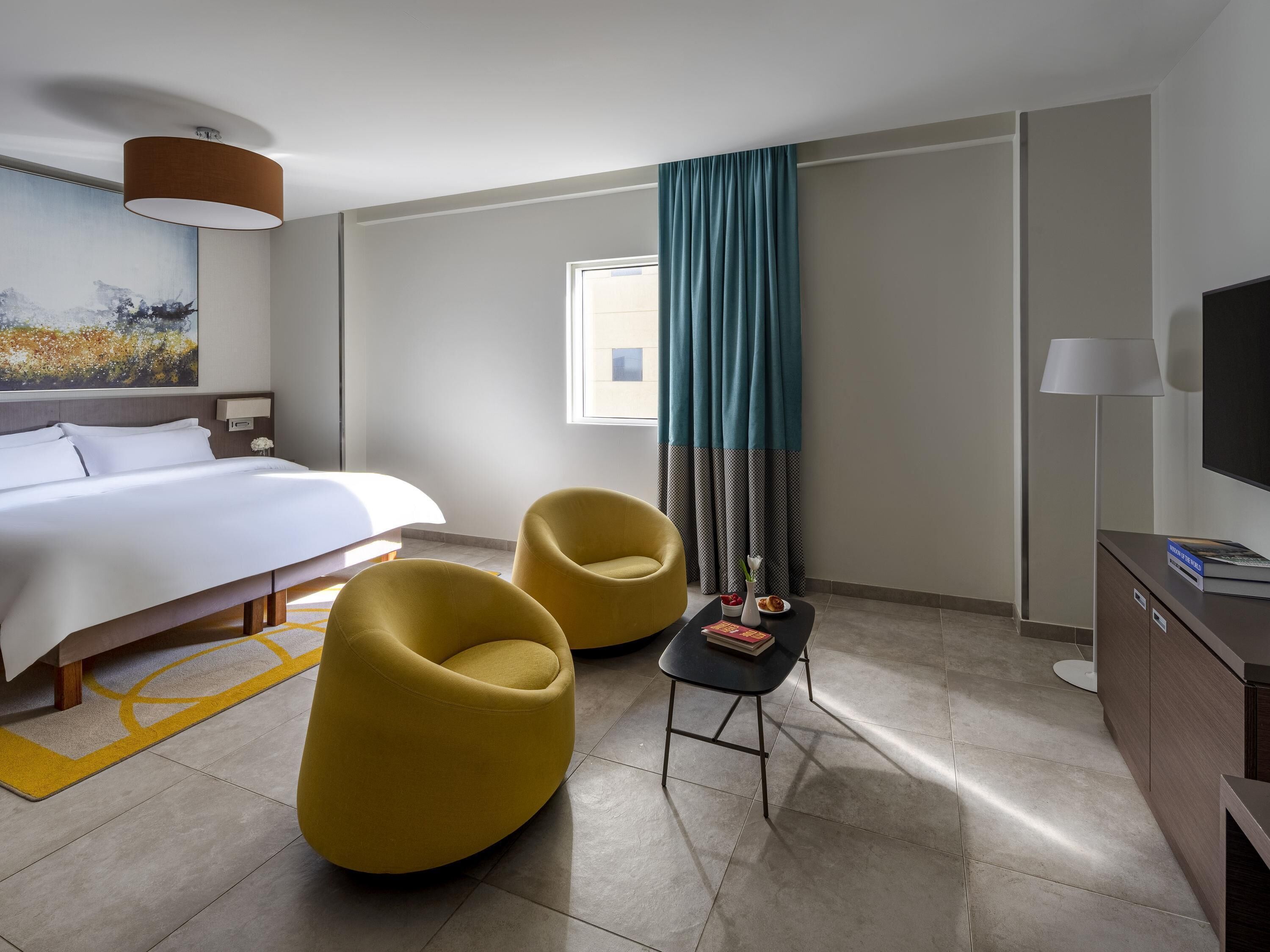 Habitación de hotel con cama, dos sillas amarillas, una mesita, televisor y ventana con cortinas.