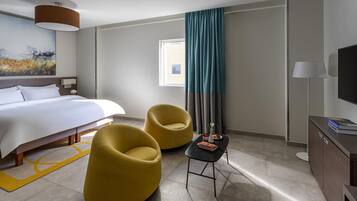 Habitación de hotel con cama, dos sillas amarillas, una mesita, televisor y ventana con cortinas.