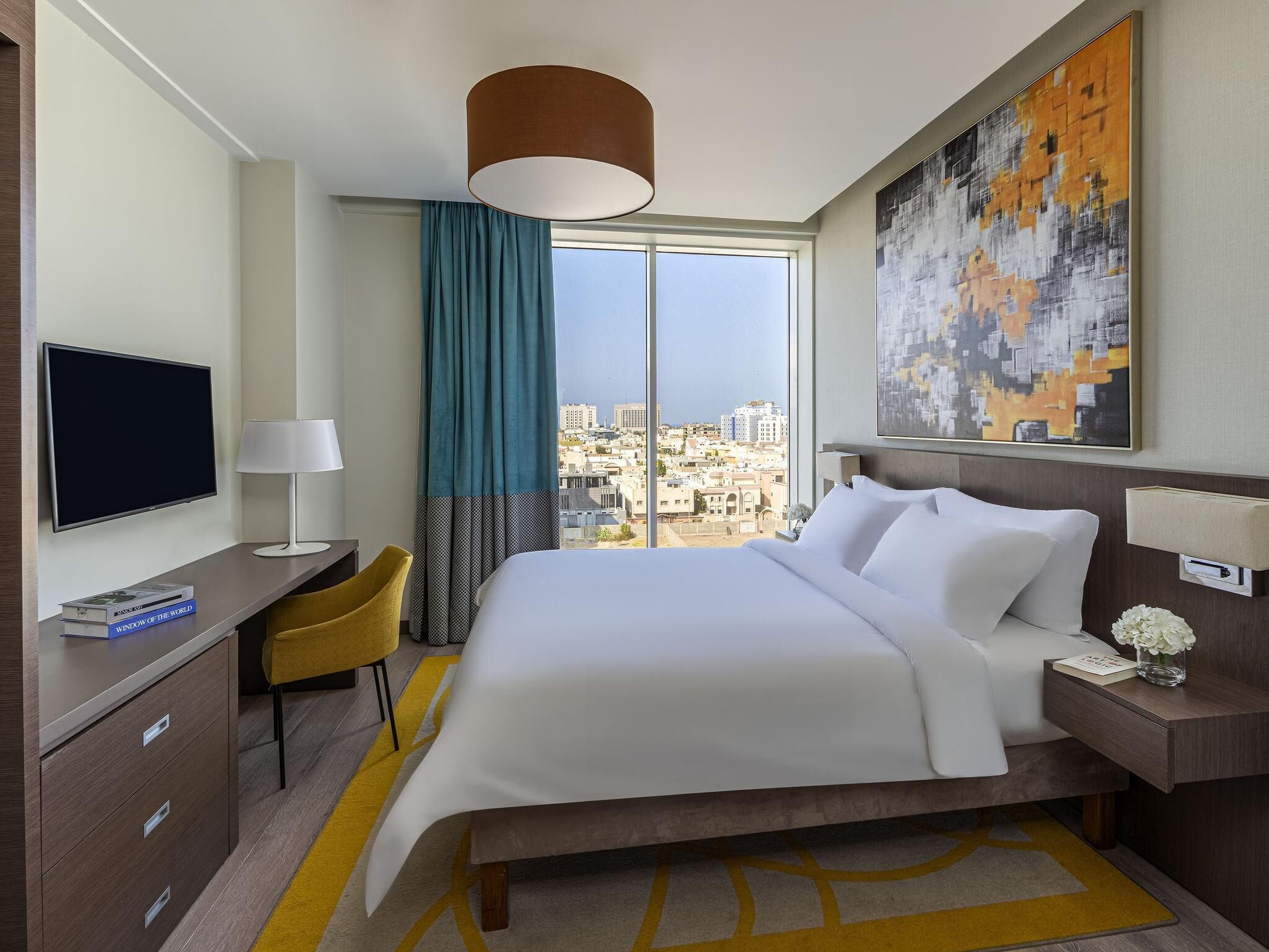 A hotel room with a bed, a desk with a TV, a chair, a window with a city view, and a painting on the wall.