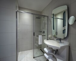 Ein modernes Badezimmer mit einer Duschkabine aus Glas, einem weißen Waschbecken mit großem Spiegel und ordentlich gefalteten Handtüchern auf einem Regal.