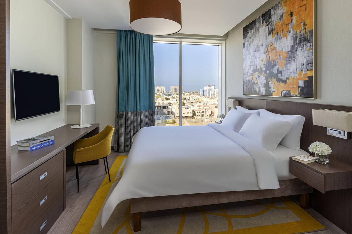 Hotelkamer met een bed, een bureau met een televisie, een stoel, een lamp, een nachtkastje met een vaas en een groot raam met uitzicht op de stad.
