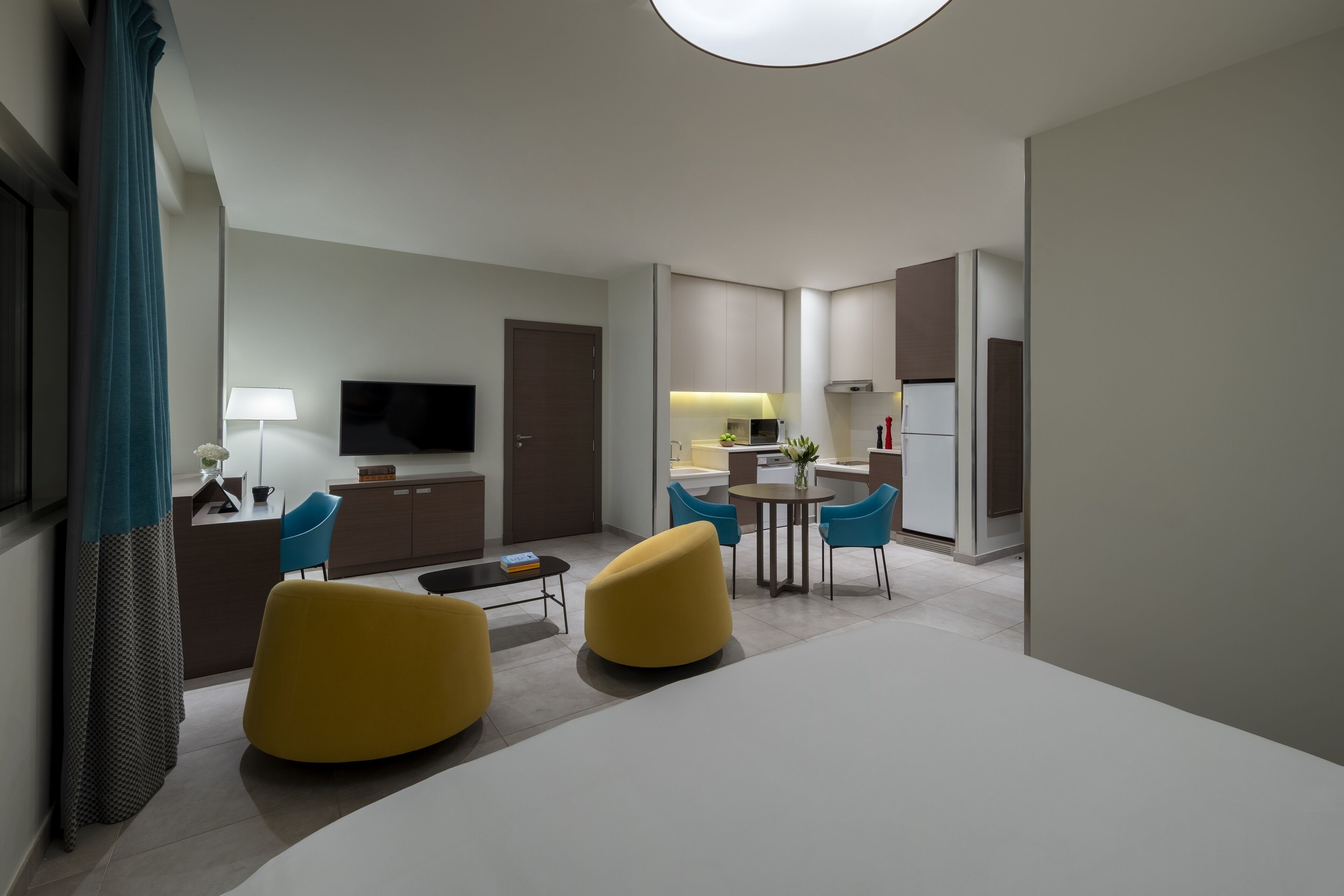 A modern hotel room with a flat-screen TV, a dining area with a round table and chairs, and a kitchen area with a refrigerator and microwave.