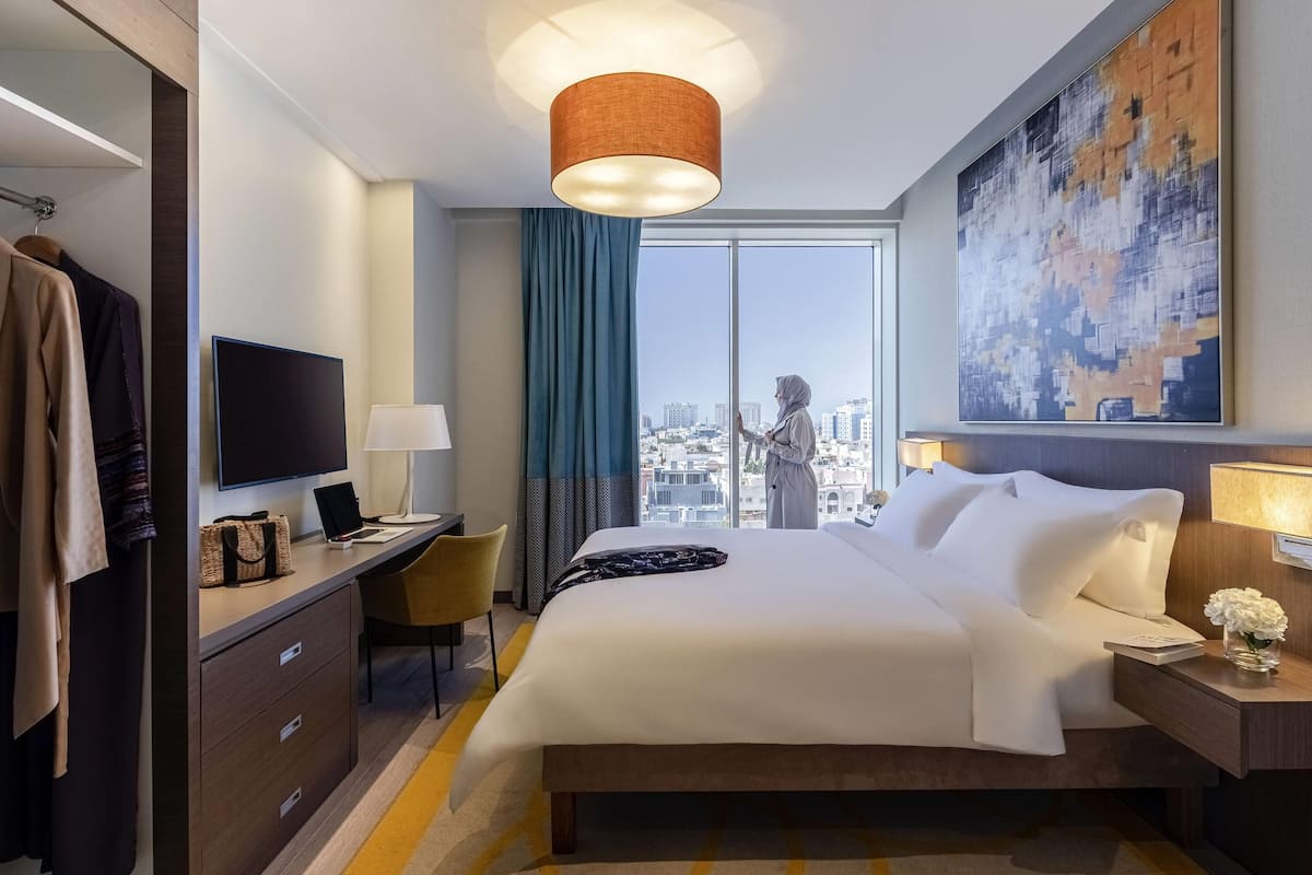 Een hotelkamer met een bed, bureau, televisie en een groot raam met uitzicht over de stad.