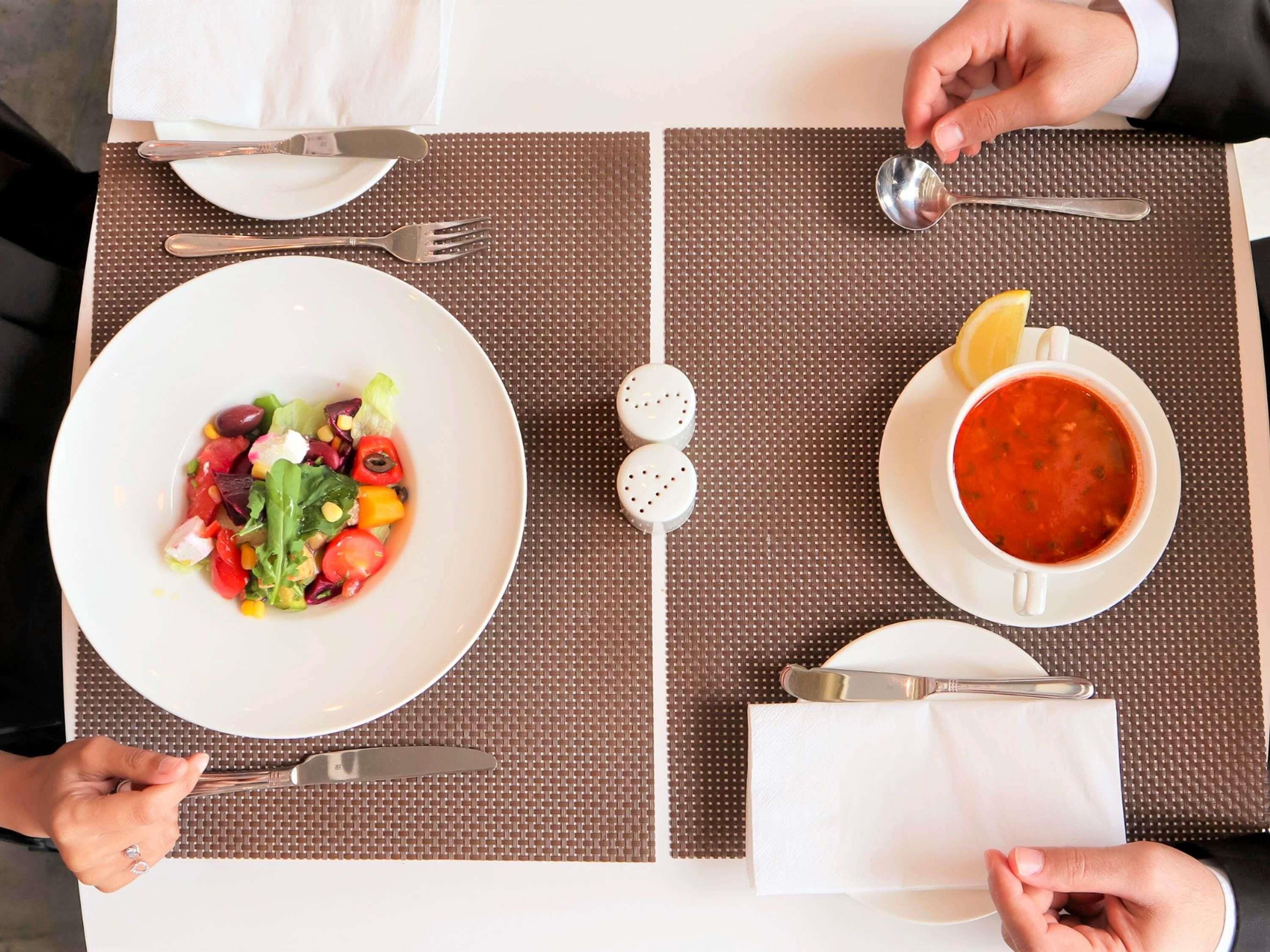 Uma mesa posta com salada, sopa e um pedaço de limão, temperos e talheres.