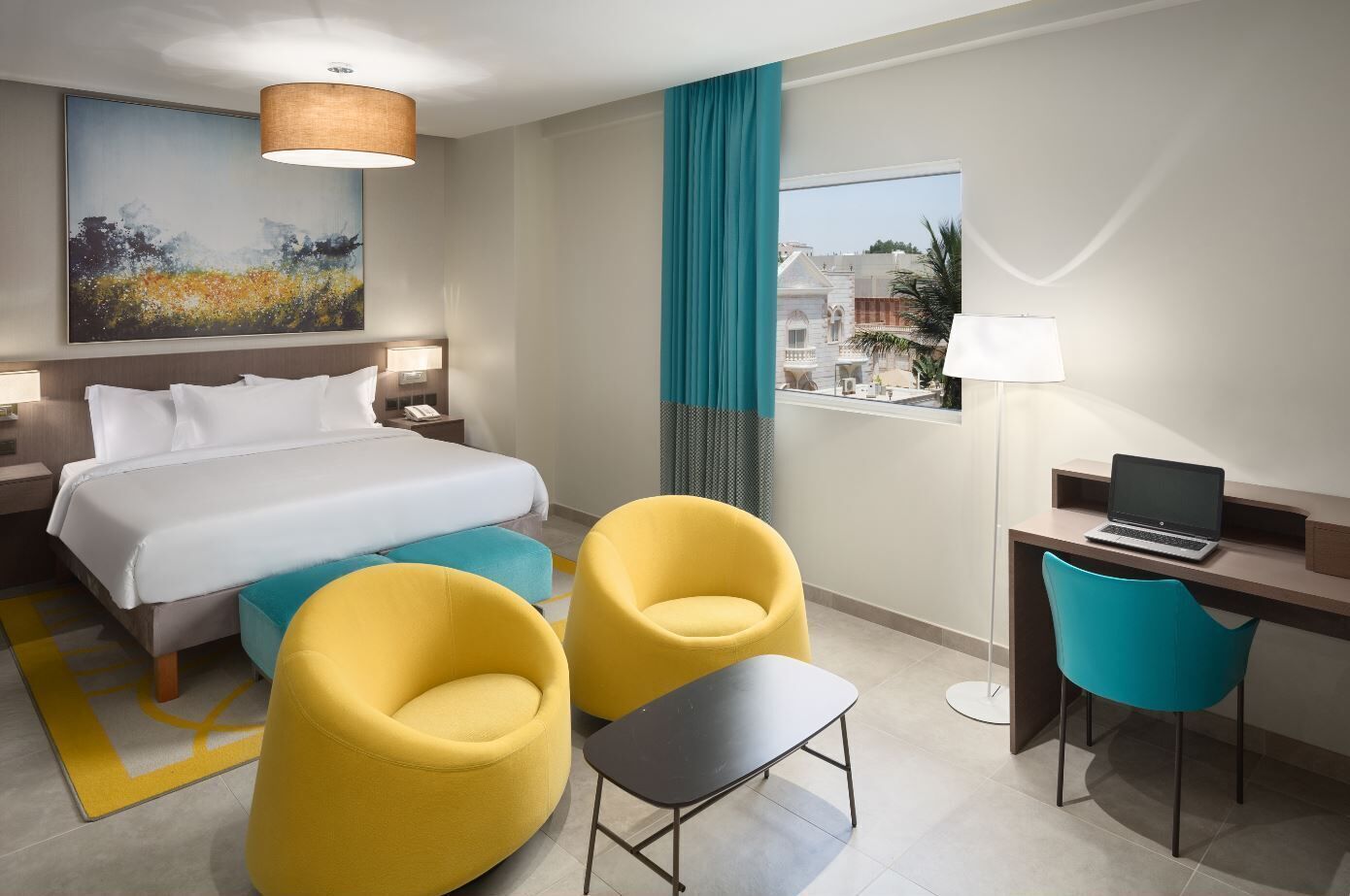 A hotel room with a bed, two yellow armchairs, a desk with a laptop, a painting on the wall, and a view of buildings outside.