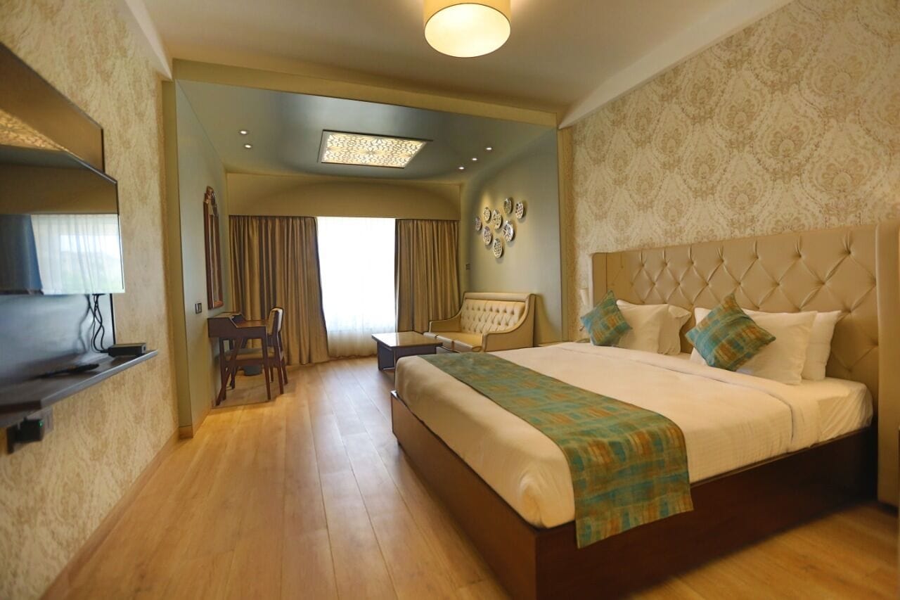 A hotel room with a large bed, a desk, and a chair. There is a window with curtains, a ceiling light, and a wall-mounted TV.