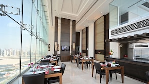A modern restaurant with a view of a city skyline, featuring high ceilings, large windows, and a mix of wooden and metal furnishings. - Harbour Suites Hotel from Holiday Gulf Hotel (Manama)