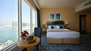 A hotel room with a large bed, a small table with fruit and flowers, a view of the city and water, and two framed pictures on the wall. - Harbour Suites Hotel from Holiday Gulf Hotel (Manama)