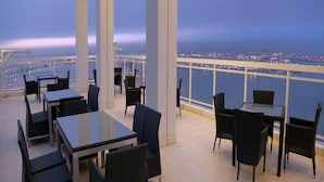 A terrace with tables and chairs, overlooking a cityscape and water at dusk. - Harbour Suites Hotel from Holiday Gulf Hotel (Manama)